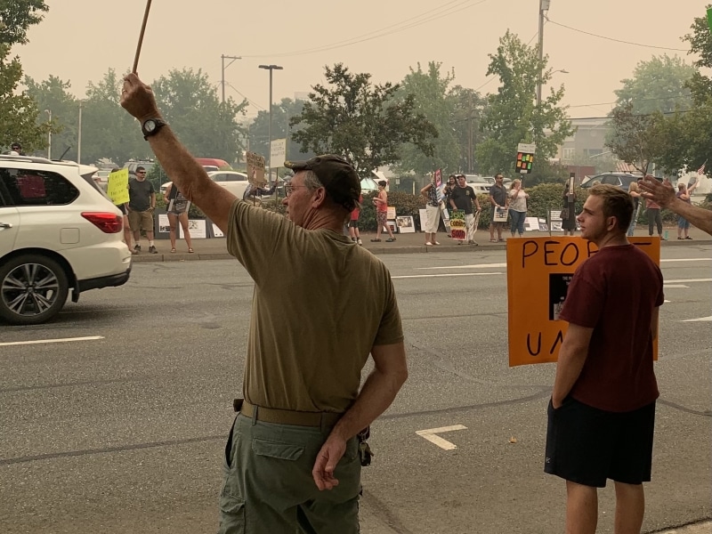 Older man stands on smoky street holding a sign raised high in the air. Sign has note in sharpie reading "Fight like hell." Another many stands nearby with an illegible sign.