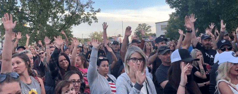 crowd of white people stand facing a speaker, with right hands raised