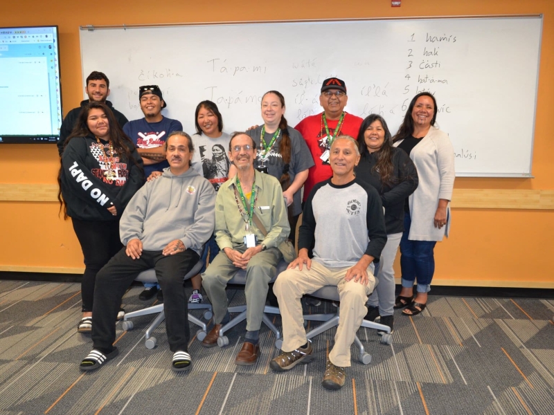 ten adults sit and stand in front of a whiteboard in a classroom, smiling at the camera