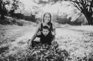 young woman with long dark hair sits outside in pile of leaves with small boy in lap