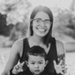 young woman with long dark hair sits outside in pile of leaves with small boy in lap