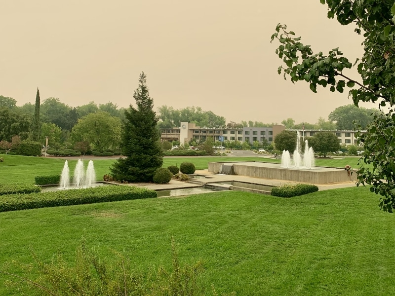 large grassy lawn with two sets of three fountains, and several trees, facing a hotel in the distance