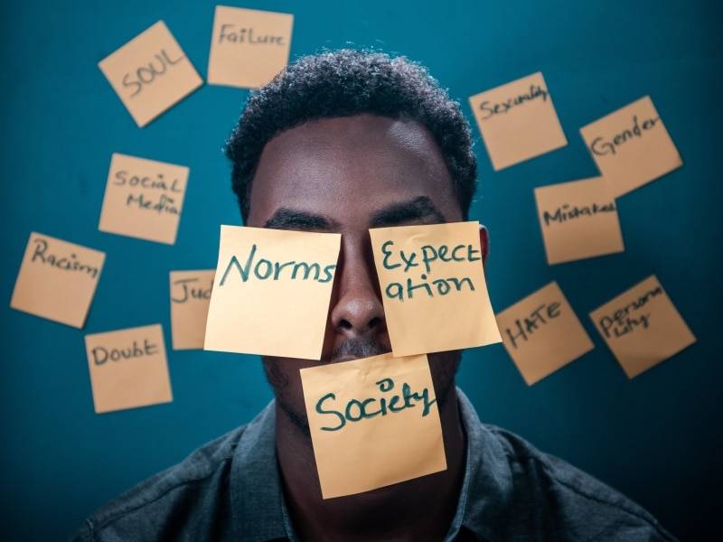 man's face against blue background mostly obscured by post it notes with words including "normal" "society" and "expectations"